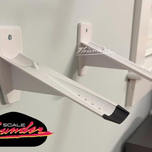 May include: Two white wall-mounted shelf brackets with a black accent on the end. The brackets are mounted on a light gray wall. The logo "SCALE Thunder" is visible in the lower left corner.