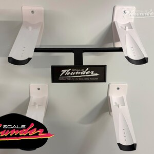 May include: White and black display shelves for model cars. The shelves are mounted on a wall and feature the "Scale Thunder" logo. The design includes four angled shelves and a central support structure, ideal for showcasing model cars.