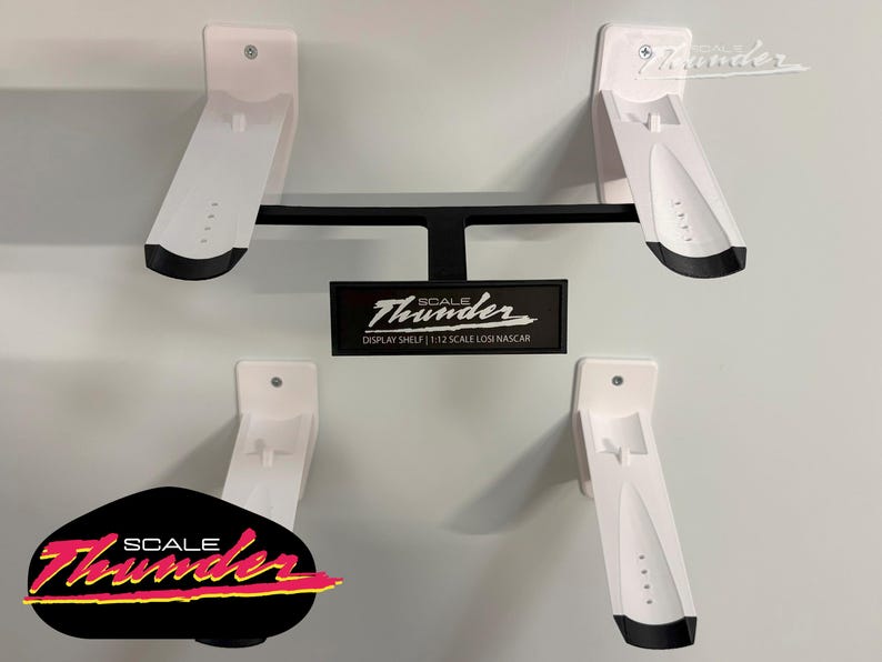 May include: White and black display shelves for model cars. The shelves are mounted on a wall and feature the "Scale Thunder" logo. The design includes four angled shelves and a central support structure, ideal for showcasing model cars.