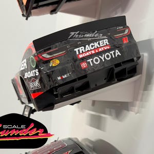 May include: A black and red toy race car with the number 19, sponsored by Tracker Boats & ATVs and Toyota. The car is displayed on a white shelf, with the "Scale Thunder" logo visible.