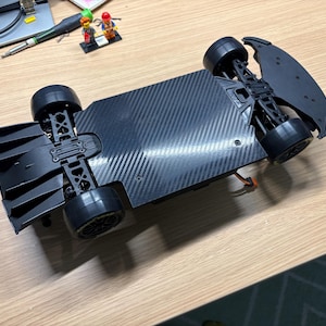 Può includere: Un telaio in fibra di carbonio nero per auto telecomandata con una base rettangolare piatta, quattro pneumatici in gomma nera, un diffusore posteriore e un paraurti anteriore. La struttura presenta componenti di sospensione meccanica.