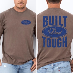 May include: Brown t-shirt with the word "Dad" in a blue oval on the front and "BUILT Dad TOUGH" on the back. The shirt is a solid brown color and has a crew neck.
