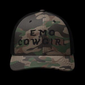 May include: A camouflage trucker hat with the words "EMO COWGIRL" in black block letters. The hat features a mesh back and a curved brim. The camouflage pattern includes shades of green, brown, and black.