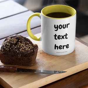 Puede incluir: Una taza de cerámica blanca con un borde y un asa amarillos. La taza está llena de café y tiene el texto "your text here" impreso en la parte delantera. La taza está sobre una tabla de cortar de madera con una pequeña hogaza de pan.