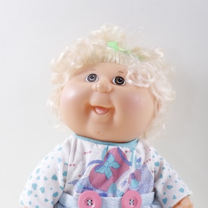 May include: A Cabbage Patch Kids doll with blonde curly hair, wearing a blue and white dress with a pink heart design and blue shoes.