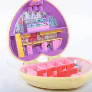 May include: A purple and yellow heart-shaped plastic toy playset with a hinged lid. The inside of the playset is divided into several compartments, each with a different animal or pet theme. The compartments include a bird cage, a fish tank, a dog kennel, and a cat bed. The playset is designed to be used as a pretend pet shop or animal shelter.