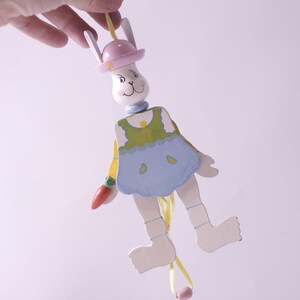 May include: A wooden pull-string toy in the shape of a bunny. The bunny has a white face, pink hat, and blue dress. It is holding a carrot and has yellow and pink strings attached to its limbs.