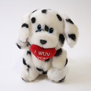 May include: A plush Dalmatian dog with black spots and blue eyes, holding a red heart-shaped pillow. The pillow has the text "I WUV YOU" in white. The stuffed animal is white with black spots.