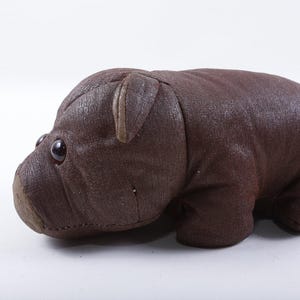 May include: A dark brown stuffed animal shaped like a hippopotamus, crafted from a textured fabric material. The figure features two small, dark, round eyes, a rounded snout, and two small ears positioned on top of its head, with visible seams.