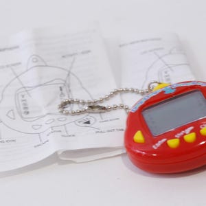 May include: Red and yellow digital pet toy with a blue and white screen. The toy has a chain attached to it and is on a white surface.