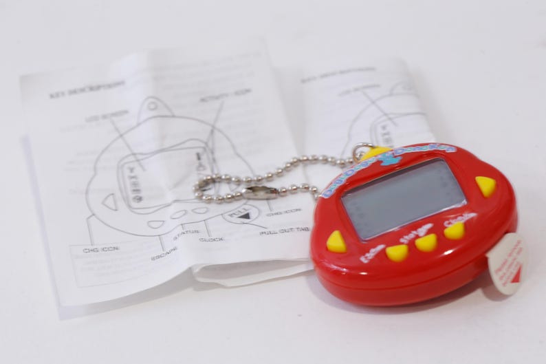May include: Red and yellow digital pet toy with a blue and white screen. The toy has a chain attached to it and is on a white surface.