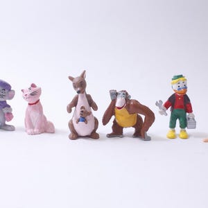 May include: A collection of small, colorful figurines of cartoon characters. The figurines include a mouse, a cat, a kangaroo with a joey, an orangutan, a man with a wrench, and a child. The figures are on a white background.