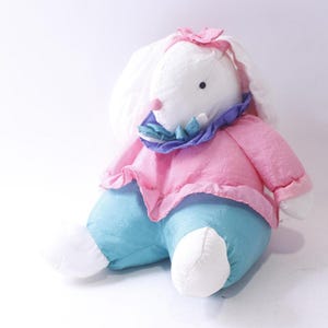 Può includere: Un peluche a forma di coniglio con pelliccia bianca, che indossa una maglia rosa a maniche lunghe, pantaloni blu acceso e un colletto arricciato viola e verde acqua. Il coniglio ha un piccolo naso rosa, occhi neri e un fiocco rosa sulla testa.