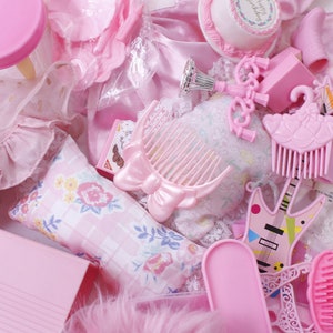 Può includere: Una collezione di giocattoli e accessori in plastica rosa per una casa delle bambole, tra cui una sedia, un pettine, una chitarra, una spazzola, un cuscino, una torta e un vestito.