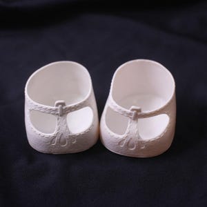 May include: A pair of white doll shoes featuring a T-strap design with molded stitching details and cut-out sections on the sides. The shoes have a rounded toe and a smooth, matte finish, designed for small figures with a low-profile silhouette.