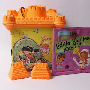 May include: Vintage orange plastic castle toy with a black handle, a yellow and green illustrated box, and a pink box with the text "Liddle Kiddles Kastle". Two orange plastic blocks are in front.