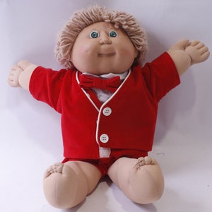 Puede incluir: Una muñeca Cabbage Patch Kid con un traje rojo y una pajarita blanca. La muñeca tiene el pelo rubio y los ojos azules.