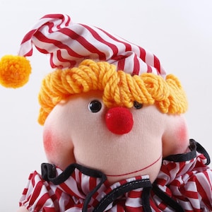 May include: A plush clown doll with a red nose, yellow yarn hair, and a red and white striped hat and collar. The doll has rosy cheeks and a stitched smile. The hat has a yellow pom-pom.