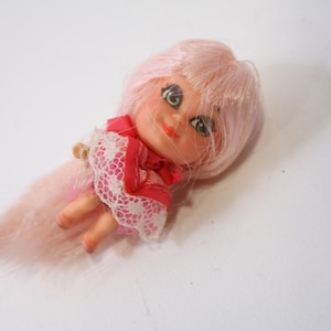 May include: A vintage doll with pink hair and a matching long tail. The doll has green eyes, a red dress with white lace trim, and small arms and legs. The doll is lying on a white surface.