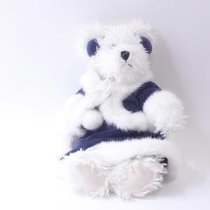 May include: White plush teddy bear wearing a blue and white fur-trimmed coat. The bear is sitting on a white background.