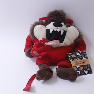 May include: A red plush devil Tazmanian Devil bean bag toy with a red pitchfork and a tag that reads "Devilish Taz Bean Bag".