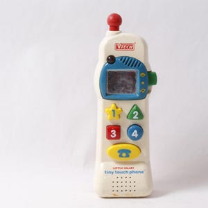 May include: A cream-colored VTech Little Smart Tiny Touch Phone toy with a red antenna, blue screen, and colorful buttons. The phone has a blue and green design with the numbers 1, 2, 3, and 4. The toy phone has the text "Little Smart tiny touch phone".