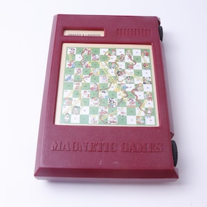May include: A red plastic travel game with a magnetic Snakes and Ladders game board. The game board has a green background with colorful illustrations of snakes and ladders. The game is labeled "Magnetic Games" and "Snakes & Ladders".