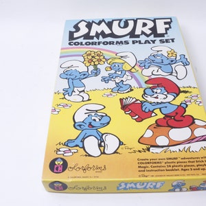 K&ouml;nnte beinhalten: Eine Smurf Colorforms Spielset-Box mit gelbem Hintergrund und bunten Smurf-Charakteren. Die Box zeigt einen Regenbogen, Blumen und einen Pilz. Der Text auf der Box lautet "Smurf Colorforms Play Set".