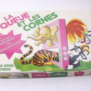May include: A vintage board game box, titled "LA QUEUE ET LES CORNES", featuring illustrations of a tiger, elephant, goat, and rooster. The box has French text and a pink starburst with text.