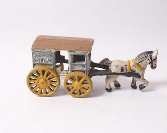 Puede incluir: Un juguete antiguo de fundición que representa un carruaje tirado por un caballo. El carruaje tiene ruedas amarillas, un techo marrón y detalles en azul y blanco. El caballo es blanco con marcas negras y un arnés amarillo.