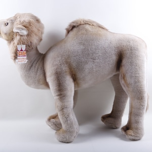 Puede incluir: Un gran juguete de peluche de camello con un pelaje marrón claro. El camello está de pie sobre sus cuatro patas y tiene un cuello largo. El juguete tiene una etiqueta que dice "Safari Original Sample".