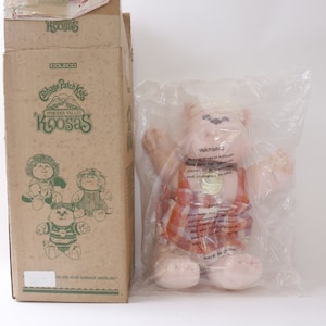 May include: A Cabbage Patch Kids Koosas doll in a red and white striped outfit with a gold medal. The doll is packaged in a clear plastic bag and comes with a cardboard box that says "Cabbage Patch Kids" and "Wisteria Valley Koosas".