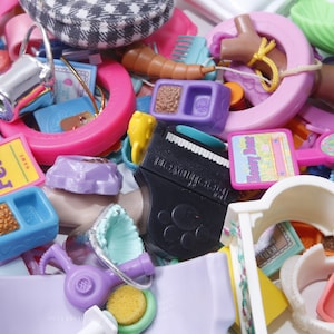 Puede incluir: Una colección de accesorios de juguete de plástico de colores para un juego de mascotas. El juego incluye una casa de juegos rosa, un tazón de comida morado, un dispensador de comida azul, un hueso amarillo, un letrero rosa y blanco que dice "Happy Hour" y un secador de pelo morado.