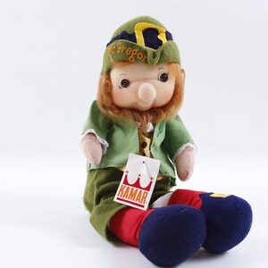 May include: A green plush leprechaun doll wearing a green hat with a gold buckle and a yellow and black striped band. The doll has a red beard, a green jacket, red pants, and blue shoes. The doll is sitting on a white surface and has a white tag with the word "Kamar" printed in red.