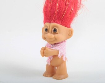 May include: A vintage Troll doll with a tan body, large eyes, and a smiling expression. It has bright red hair with gold tinsel and wears a pink and white checkered scarf. The doll is standing upright.