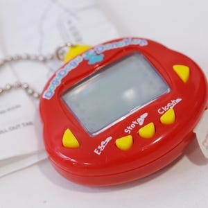 May include: Red plastic toy with a digital screen and buttons labelled "Start", "Esc", and "Clock". The toy has a chain attached and says "Dog Dayz Digimon" on the front.