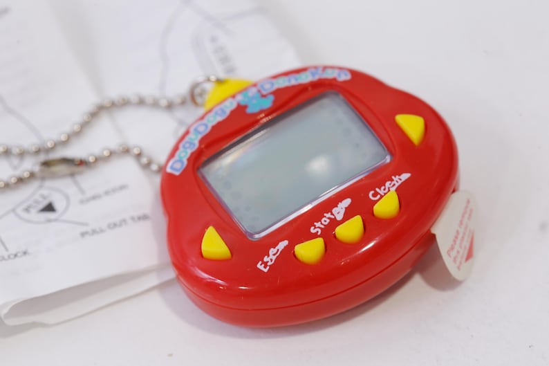 May include: Red plastic toy with a digital screen and buttons labelled "Start", "Esc", and "Clock". The toy has a chain attached and says "Dog Dayz Digimon" on the front.