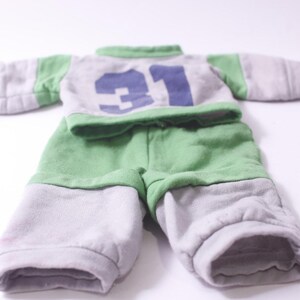 May include: A two-piece doll outfit in gray and green. The top has green sleeves and collar, with the number 31 in blue on the front. The shorts are green and gray. The outfit is made of soft fabric.