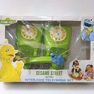 Puede incluir: Un juego de teléfono intercomunicador de pilas de Sesame Street en una caja verde y amarilla. La caja presenta imágenes de Big Bird, Cookie Monster y dos niños usando los teléfonos. El texto en la caja dice "Sesame Street Battery Inter-Com Telephone Set" y "Para edades de 3 años en adelante".