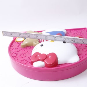 May include: Pink Hello Kitty makeup compact with a white Hello Kitty face, a pink bow, and a brown bear with a blue hat. The compact is 11 inches wide.