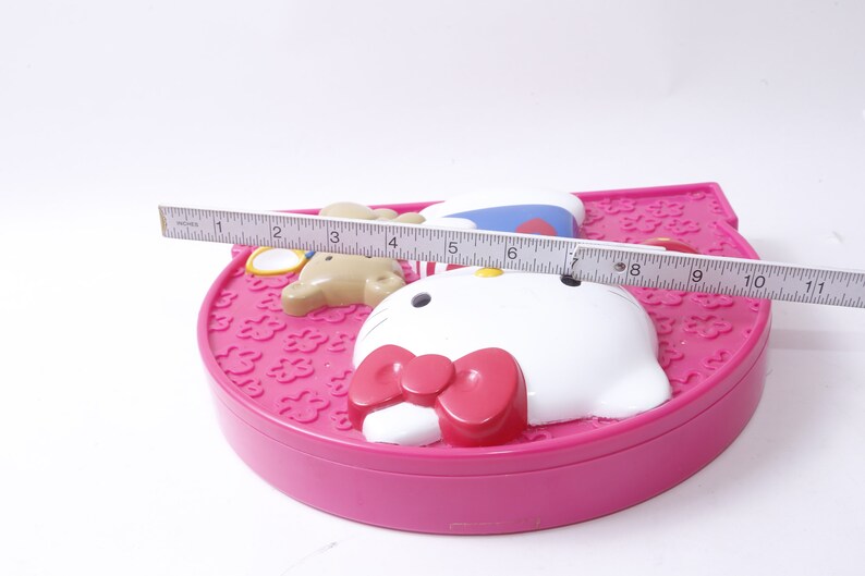 May include: Pink Hello Kitty makeup compact with a white Hello Kitty face, a pink bow, and a brown bear with a blue hat. The compact is 11 inches wide.