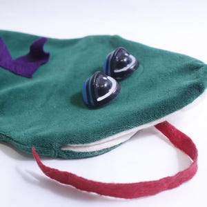 May include: A green fabric creature toy featuring a purple felt spine, two dark blue and white rounded plastic eyes, and a long red felt tongue extending from a white fabric mouth opening. The object has a soft, plush-like texture throughout.