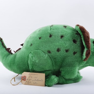 May include: Green plush toy dinosaur with brown accents. The dinosaur has three horns on its head and a spiked tail. The toy is labeled "Dinosaurs FS-111 3/2 wholesale 66¢ UK."