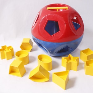 May include: A red and blue plastic shape-sorting ball with a yellow handle on top, featuring various geometric cutouts. Surrounding the ball are eleven yellow plastic shapes, including a star, a cross, a cylinder, and various polygons.