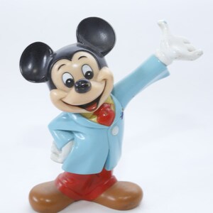 May include: A blue and red plastic figurine of Mickey Mouse wearing a blue suit with a red bow tie. He is smiling and waving with his right hand.