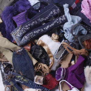 Peut inclure: Une collection de vêtements et d'accessoires pour poupées, comprenant des pantalons en jean, des sacs à main et des hauts de différentes couleurs comme le bleu, le violet, le rose et le beige. Certains articles ont des ornements décoratifs.