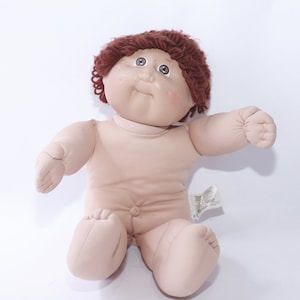Può includere: Una bambola Cabbage Patch Kid con capelli di lana marroni, seduta con le braccia distese. La bambola ha un'incarnato marrone chiaro e indossa una camicia bianca.
