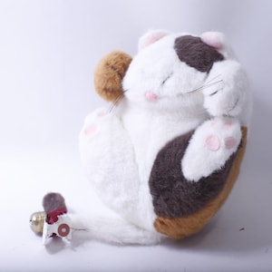 May include: A white, brown, and tan plush cat toy with a pink nose and paw pads. The cat is curled up in a sleeping position.