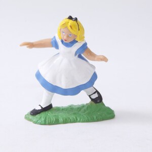 May include: A plastic figurine of Alice from Alice in Wonderland. She is wearing a blue and white dress and black shoes. She is standing on a green base.