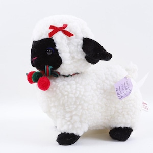 May include: A plush lamb toy with white curly fur, a black face, and black ears. It features a red ribbon bow on its head and a collar with red and green pom-poms. A small purple tag is attached to its side, and it stands on four black hooves.
